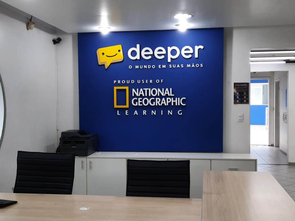 Parte interior do prédio da escola de idiomas, onde aparece uma parede azul com o logo da escola e o logo do National Geographic