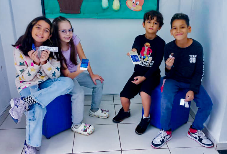 Quatro alunos de cerca de 8 anos de idade, sentados exibindo seus trabalhos artísticos realizados durante as aulas